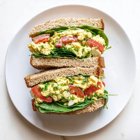 Avocado egg salad sandwich on whole grain bread, garnished with chives and served with a side of mixed greens.  