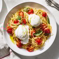Creamy burrata melting into warm spaghetti with juicy cherry tomatoes and fresh basil leaves on a white plate.